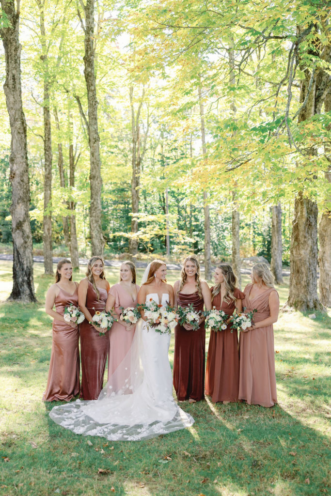 bride and bridesmaids standing in a maple grove wearing mismatched pink dresses