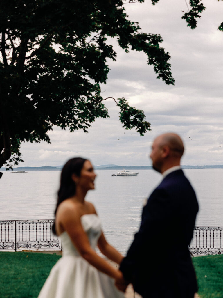 Bride and groom hold hands on a lawn overlooking the ocean--they are blurred while the focus of the image lands on a white yacht floating in the harbor beyond them.