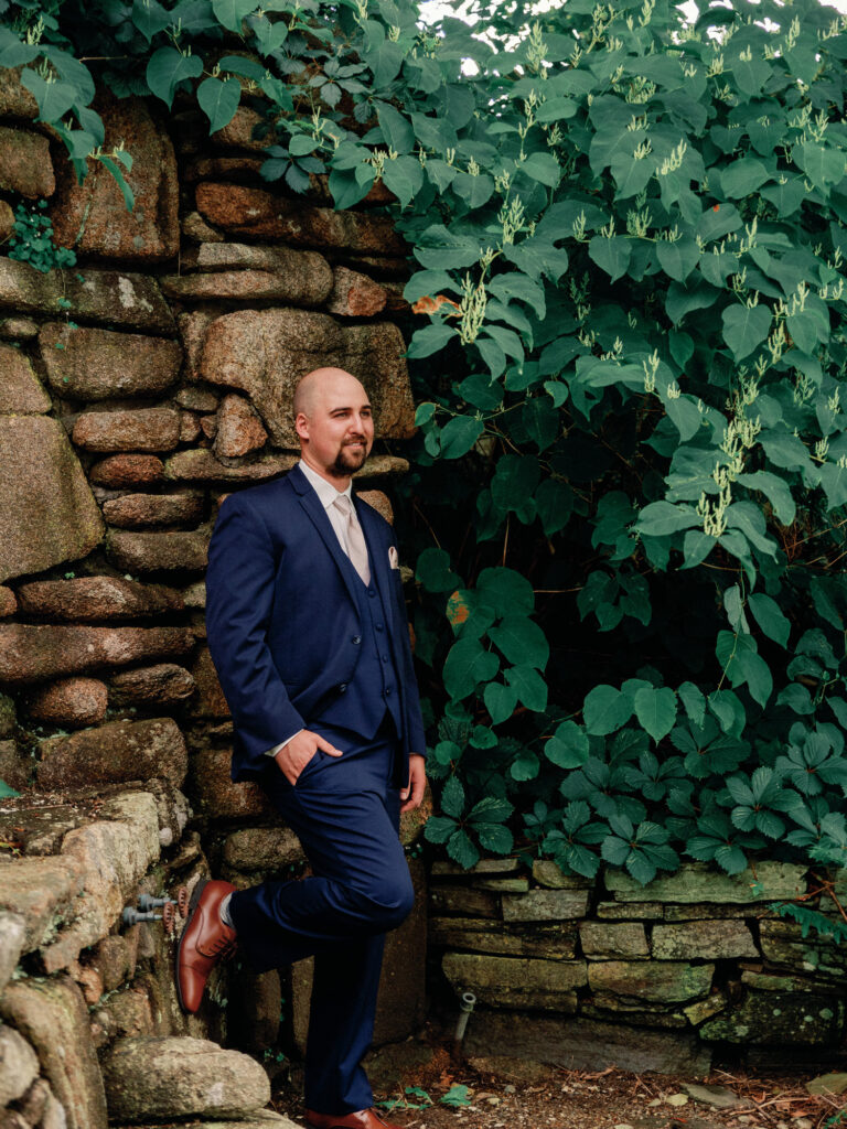 Groom poses for a relaxed portrait while leaning on a stone wall covered in ivy.