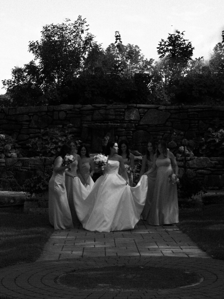 Reportage, The Friends Club inspired image of a Bride and her bridesmaids who carry her dress walking through a streak of light in a black and white wedding photo.