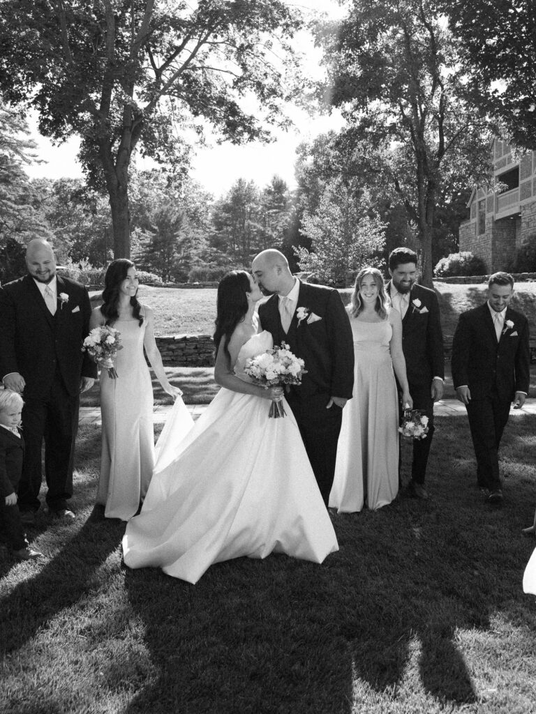 Black and white portrait of the wedding party walking alongside the bride and groom as they share a kiss.