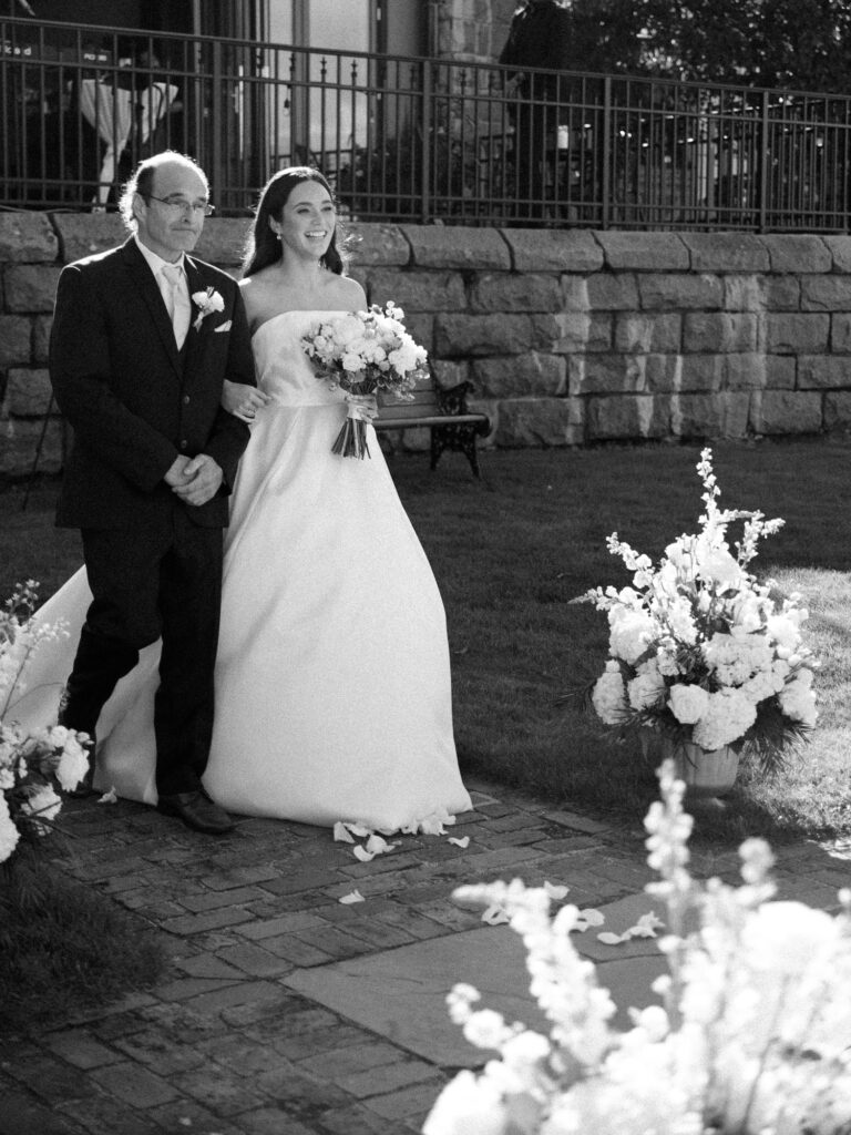 Bride and her father step into the aisle as they begin to walk toward the wedding ceremony, in black and white.