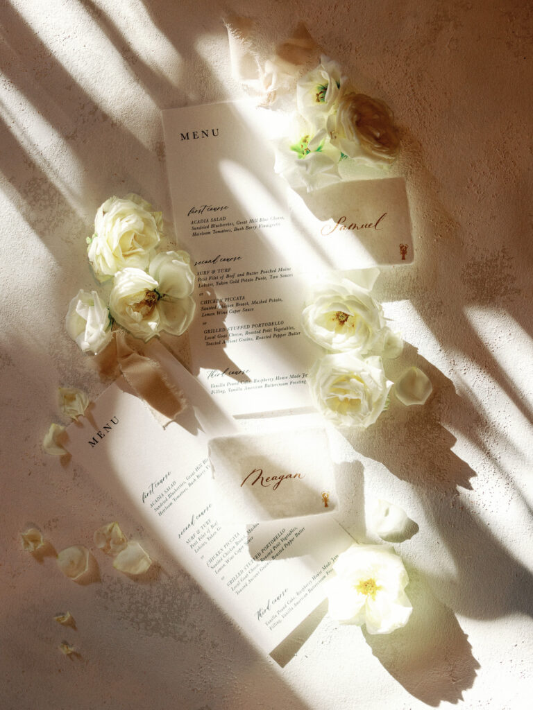 Glowing portrait of hand-crafted wedding stationery (gold-ink name cards and custom menus) surrounded by flowers and dappled streaks of light and shadow in an incredible flat lay by Maine wedding photographer Kay Cushman.