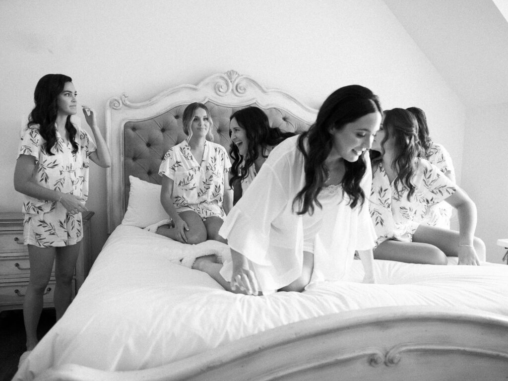 Photojournalistic view of bride and bridesmaids getting on a bed for a portrait together while wearing matching pajamas on a wedding day