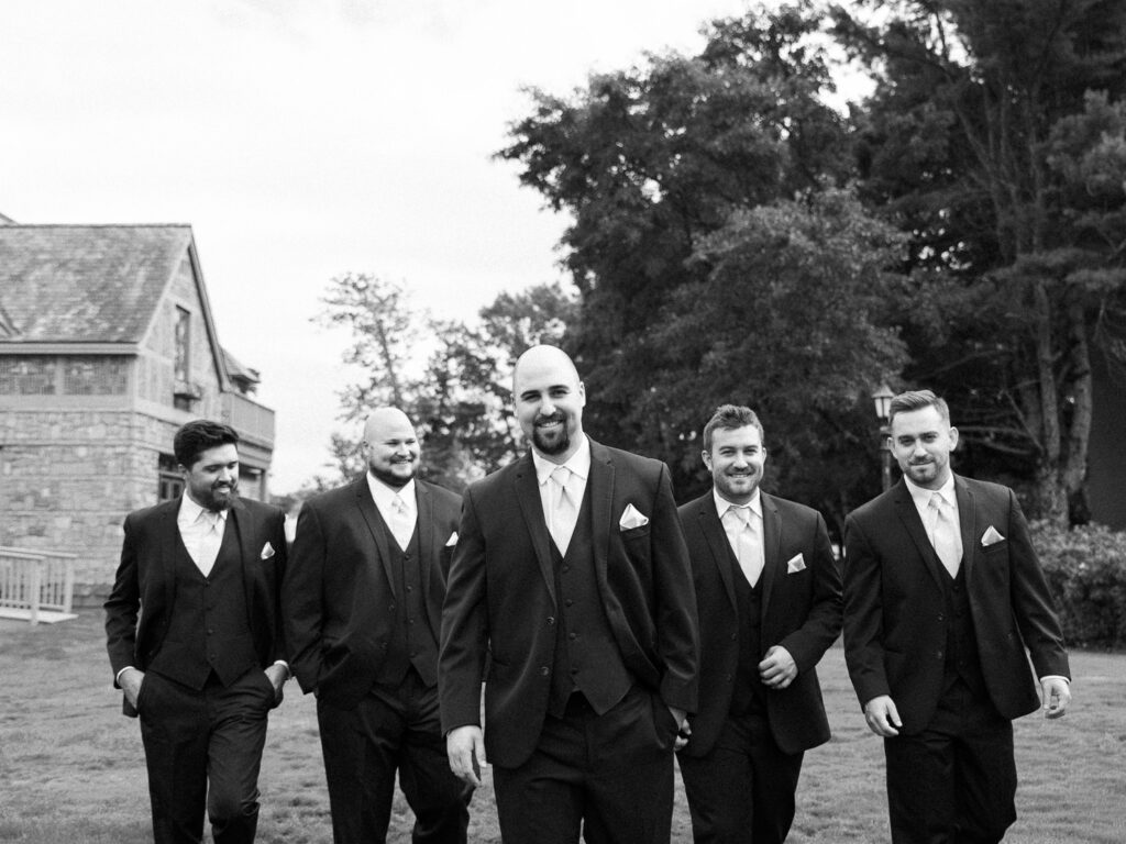 Groom walking in the center of four groomsmen on a wedding day while wearing navy blue tuxes.