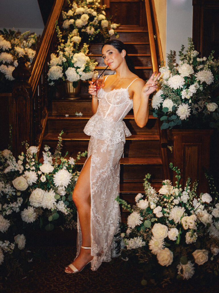 Bride throws a peace sign while drinking a pink cocktail at her wedding reception. She poses in front of a large staircase covered in flowers while wearing a lace dress. Cool girl chic vibes like The Friend Club, a wedding blog that shares trendy and emotional wedding content.