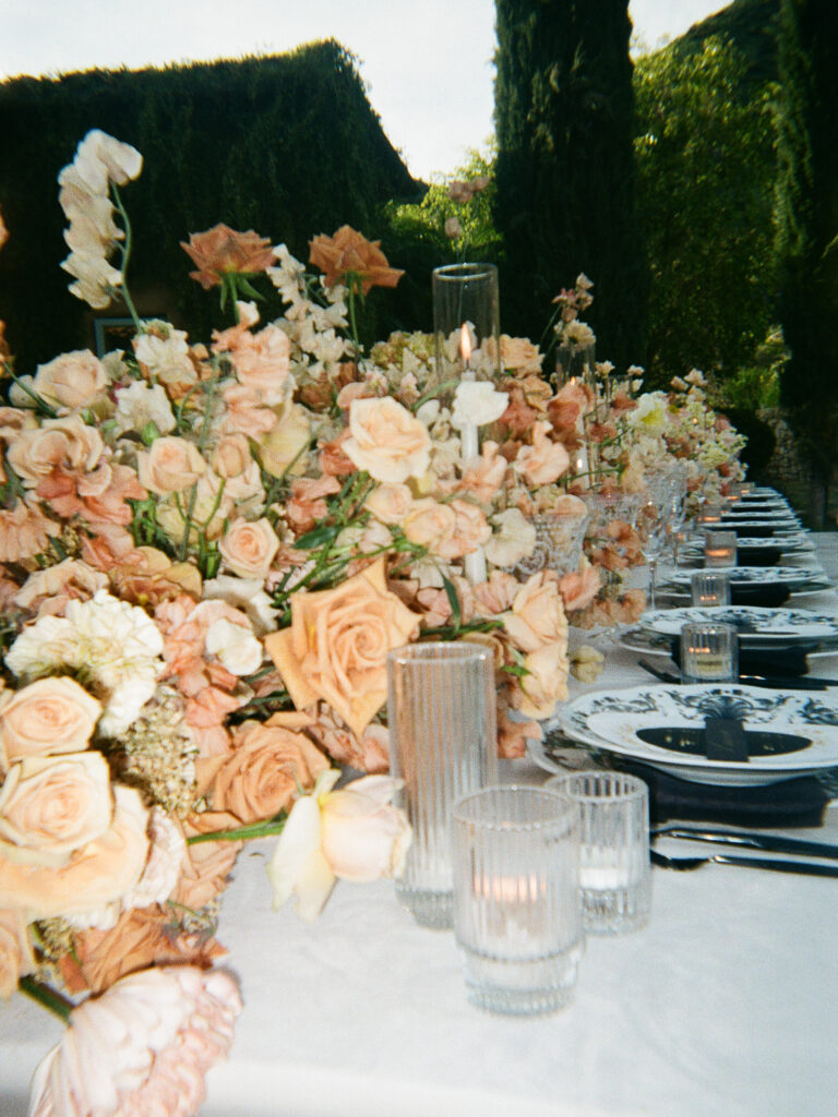 Roses, poppies, sweat peas, and ranunculus form a terracotta floral arrangement by Creative Light Design spanning the length of a table in a wedding photo in California.