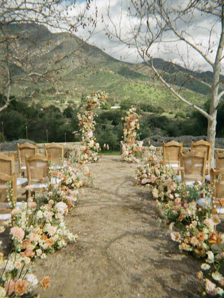 Aisle decorated with terracotta and pink flowers flank wicker-backed chairs at a luxury wedding wedding in Ojai. The mountain views behind the asymmetrical flower arch complete the look. Flowers crafted by Creative Light Design, a wedding florist in Southern California.