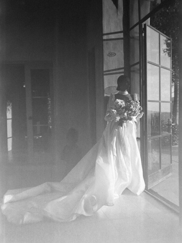 Bride in a flowing Leah da Gloria wedding dress rests by a window in black and white film wedding photos with a bouquet from Creative Light Design.