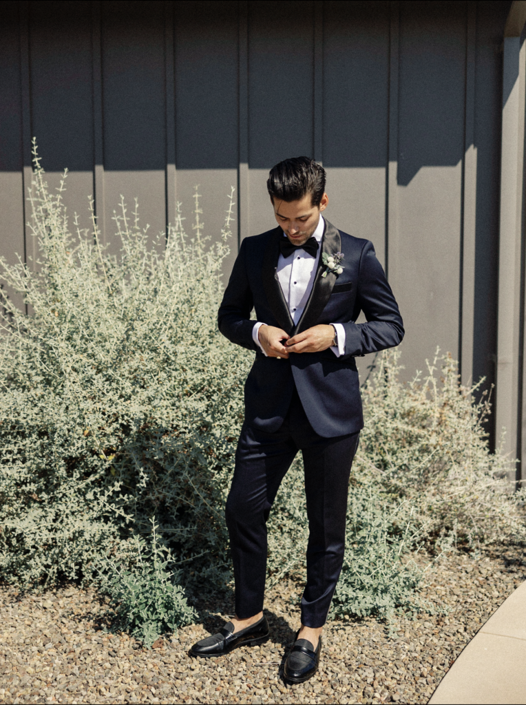 Groom in navy blue tux with black lapels stands in the sunshine outside a suite at Stanley Ranch during his wedding in Napa.