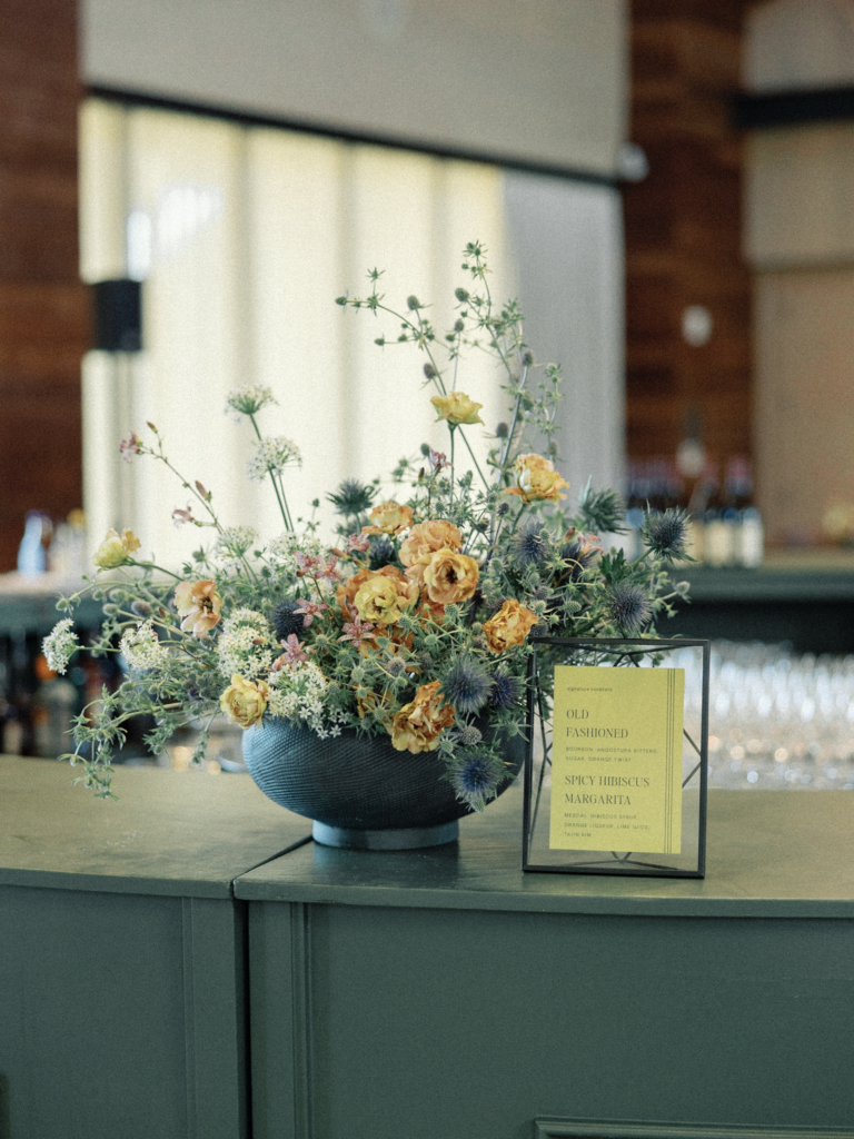 Large vase of orange, blue, and white wild flowers sits on a bar during a wedding's cocktail hour.