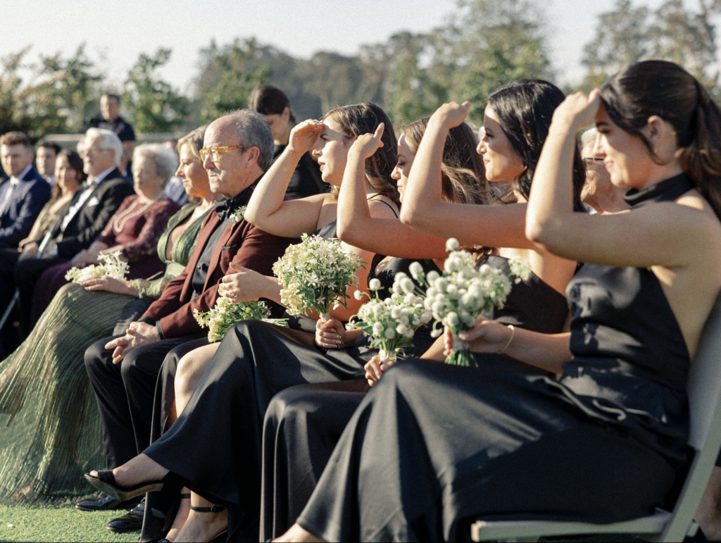 Bridesmaids wearing black dresses with white bouquets sit in the direct sun and shield their eyes during a luxury wedding ceremony in Napa, California.