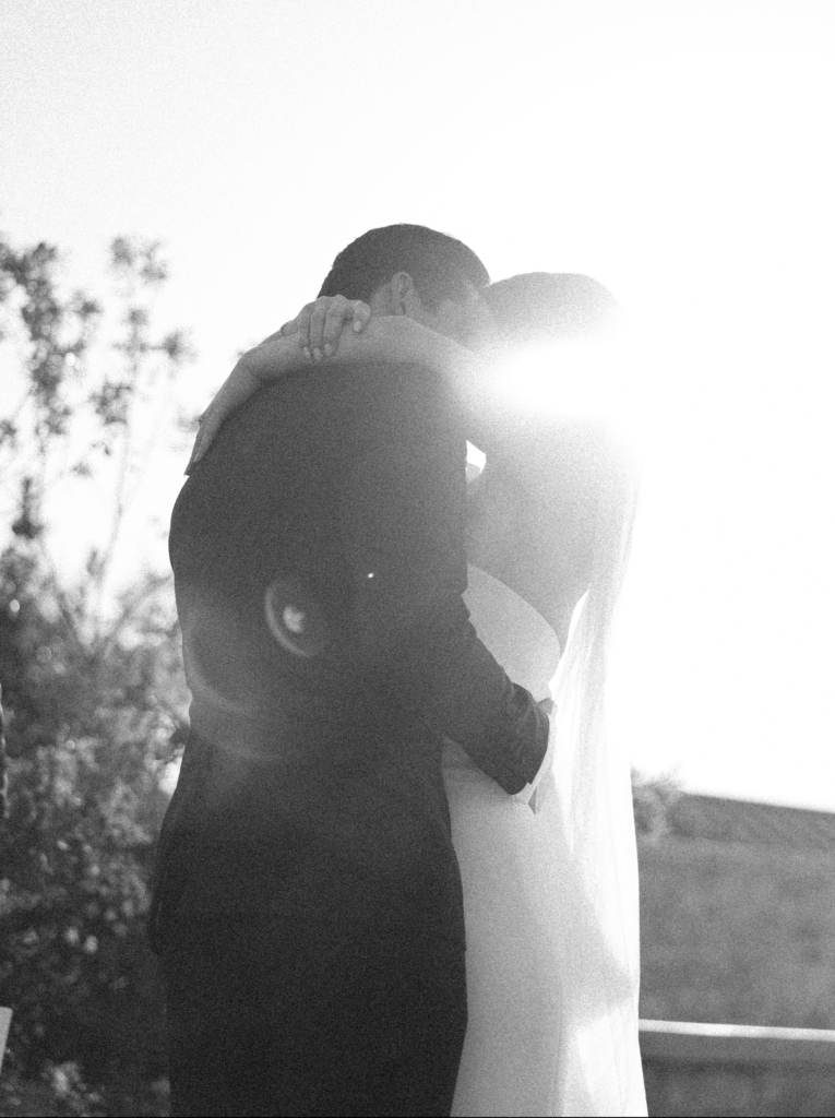 Black and white wedding film photo of bride and groom kissing in the sun while hugging in a romantic pose.