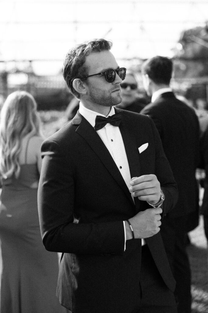 Man in a black tuxedo fixes his watch while waiting for a drink at a wedding's cocktail hour.