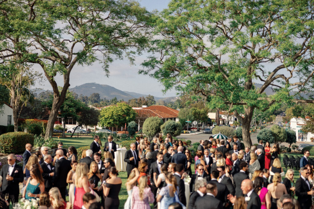 Guests mingle on the lawn of the Inn at Rancho Santa Fe during cocktail hour of a black tie wedding. They are backed by historic trees and rolling hills.