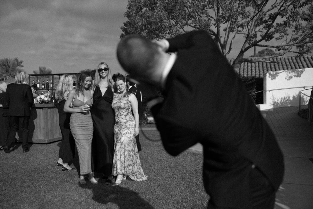 A mean leans in front of a photographer to take a picture of three women on his phone during a black tie wedding in SoCal.