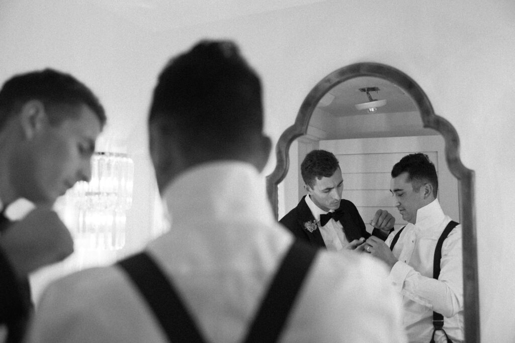 A layered image of a groom receiving help with his cufflinks as reflected by the mirror the groom and a groomsman are standing in front of.
